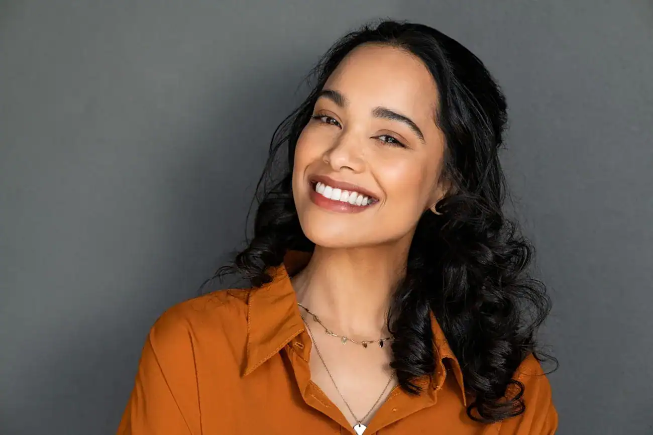 Woman with curly black hair wearing an orange shirt and layered necklaces against a gray background